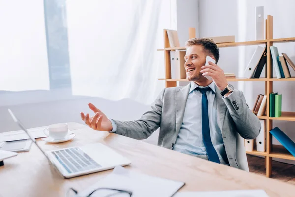 Homme d'affaires positif parlant sur smartphone près d'un ordinateur portable et café au premier plan flou au bureau — Photo de stock