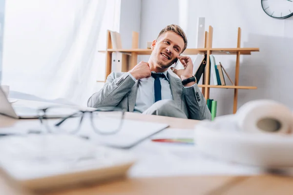 Empresário sorridente falando no smartphone perto de óculos e papéis em primeiro plano desfocado no escritório — Fotografia de Stock