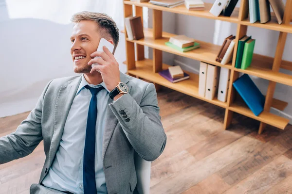 Young businessman in suit smiling while talking on smartphone in office — Stock Photo