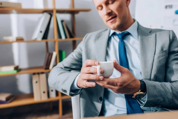 Cropped view of cup of coffee in hands of businessman on blurred background in office — Stock Photo