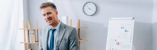 Молодой бизнесмен в костюме смотрит в камеру рядом с флипчартом на размытом фоне в офисе, баннер — стоковое фото