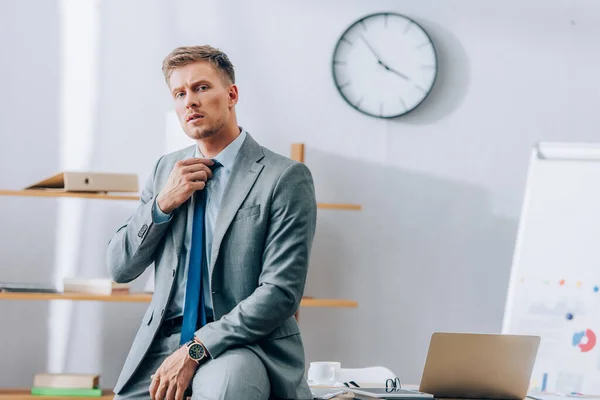 Empresario en traje ajustando corbata cerca de laptop y anteojos en mesa - foto de stock