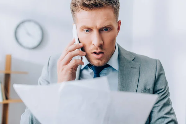 Excited businessman talking on smartphone and holding papers on blurred foreground — Stock Photo