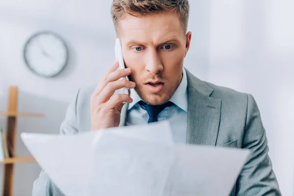 Aufgeregter Geschäftsmann spricht auf Smartphone und hält Papiere im verschwommenen Vordergrund — Stockfoto