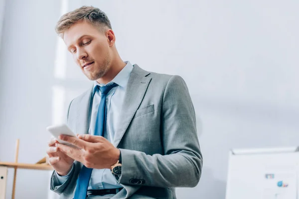 Young businessman in formal wear using smartphone on blurred foreground in office — Stock Photo