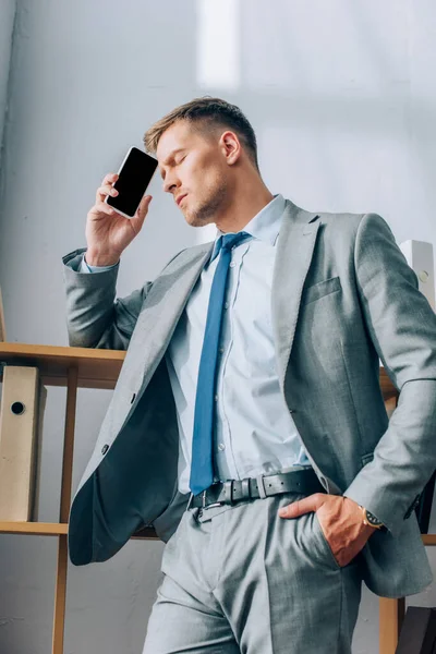 Hombre de negocios cansado sosteniendo teléfono inteligente con pantalla en blanco en la oficina - foto de stock