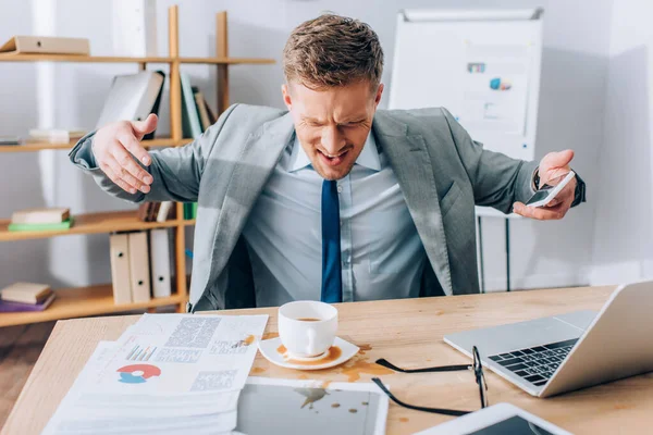 Разгневанный бизнесмен со смартфоном смотрит, как разливают кофе на газеты — стоковое фото