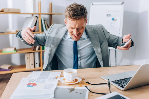 Homme d'affaires en colère avec smartphone qui cherche à verser du café sur les papiers — Photo de stock