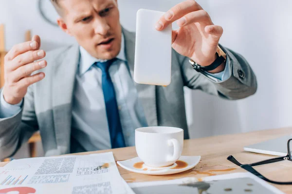 Empresário confuso segurando smartphone sujo perto derramando café e papéis na mesa — Fotografia de Stock