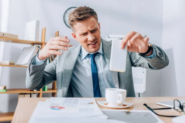 Uomo d'affari confuso che tiene lo smartphone sporco vicino a versare il caffè su carte e tavolo — Foto stock