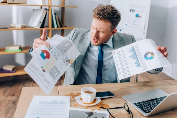 Aggressive businessman holding dirty documents near pouring out coffee and devices in office — Stock Photo