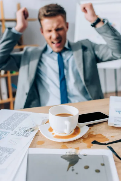 Выливание кофе на смартфон и бумаги на грязный стол рядом с сердитым бизнесменом на размытом фоне — стоковое фото
