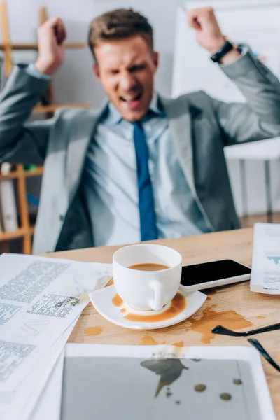 Despejando café no smartphone e papéis na mesa suja perto de empresário irritado no fundo turvo — Fotografia de Stock