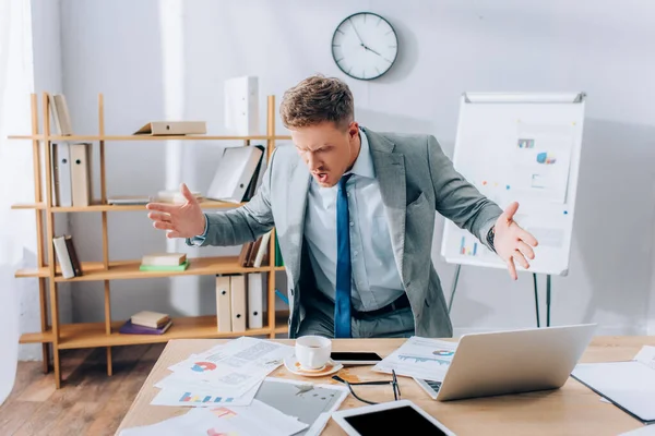 Angry businessman looking at pouring out coffee on documents near devices on table — Stock Photo