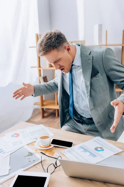 Irradiated businessman standing near pouring out coffee on documents and smartphone in office — Stock Photo