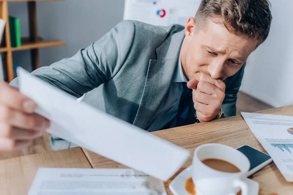 Displeased businessman holding document near pouring out coffee on blurred foreground in office — Stock Photo