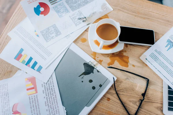 Vista superior de derramar café perto de papéis bagunçados, óculos e smartphone na mesa — Fotografia de Stock