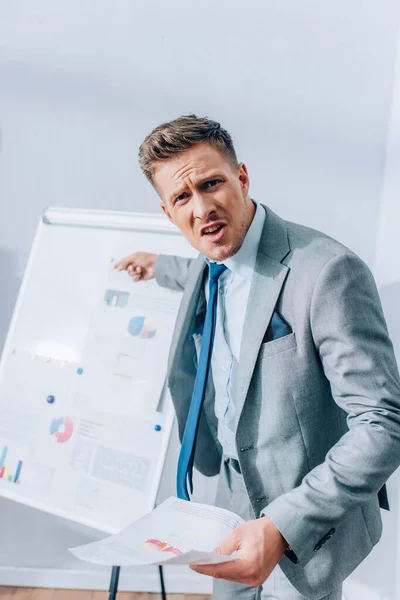 Wütender Geschäftsmann blickt in Kamera und zeigt auf Flipchart mit Grafiken auf verschwommenem Hintergrund im Büro — Stockfoto