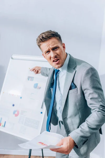 Homme d'affaires en colère regardant la caméra tout en pointant vers le tableau à feuilles avec des graphiques sur fond flou dans le bureau — Photo de stock