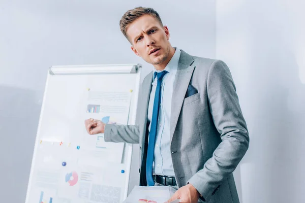 Konzentrierter Geschäftsmann blickt in die Kamera und zeigt auf Flipchart auf verschwommenem Hintergrund im Büro — Stockfoto