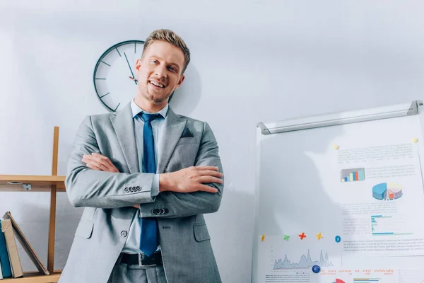 Smiling businessman with crossed arms looking at camera near flipchart with graphs — Stock Photo