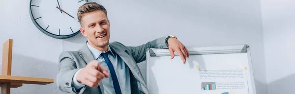 Positive businessman pointing with finger at camera near flipchart with graphs, banner — Stock Photo
