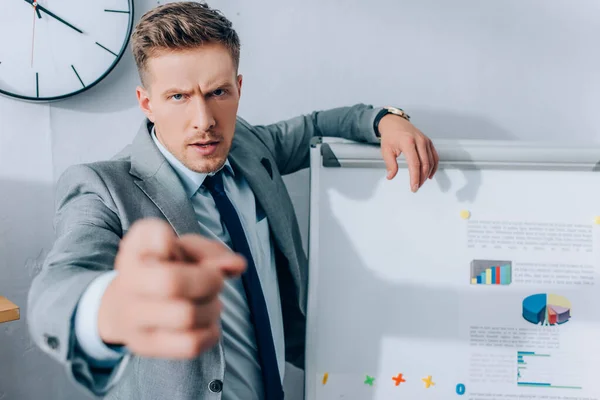 Hombre de negocios serio apuntando con el dedo a la cámara en primer plano borroso cerca del rotafolio con gráficos - foto de stock