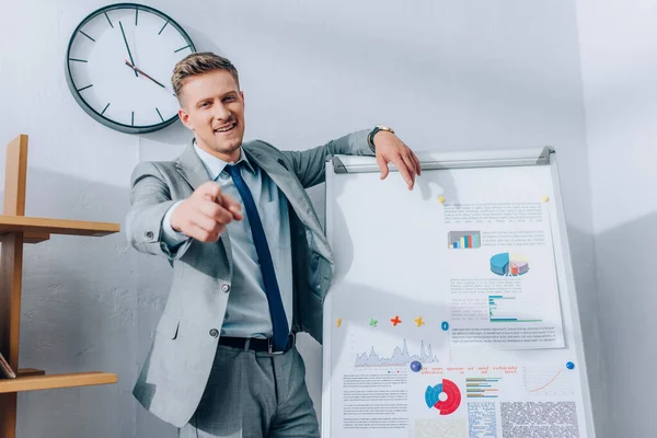 Cheerful businessman pointing at camera while standing near flipchart with graphs — Stock Photo