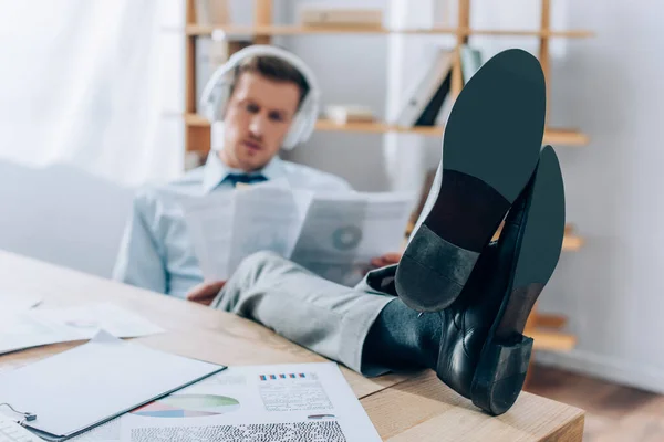 Uomo d'affari con gambe sul tavolo utilizzando cuffie e lavorando con documenti su sfondo sfocato in ufficio — Foto stock