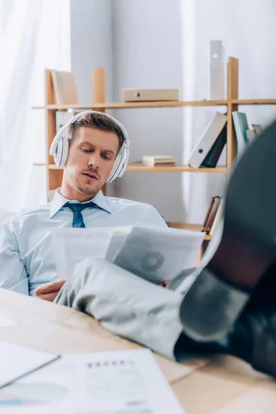 Businessman in headphones working with papers while putting legs on table on blurred foreground — Stock Photo