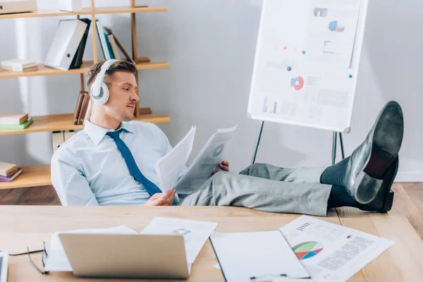 Jeune homme d'affaires dans les écouteurs travaillant avec des documents près d'un ordinateur portable sur le premier plan flou — Photo de stock