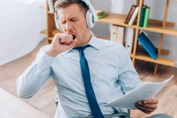 Jovem empresário em fones de ouvido bocejando enquanto mantém documentos no escritório — Fotografia de Stock