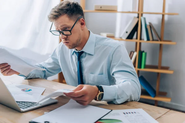 Hombre de negocios cansado sosteniendo documentos cerca de la computadora portátil en primer plano borroso en la oficina - foto de stock