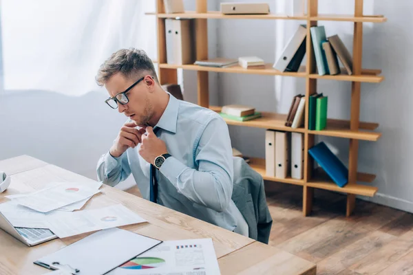 Businessman in eyeglasses touching tie while looking at paperwork near laptop on blurred foreground — Stock Photo