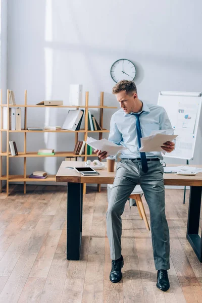 Concentrated businessman looking at papers near coffee to go and digital tablet on table — Stock Photo