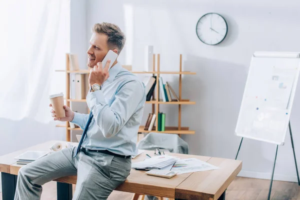 Uomo d'affari positivo che parla su smartphone mentre tiene in mano il caffè per avvicinarsi al tavolo da lavoro in ufficio — Foto stock