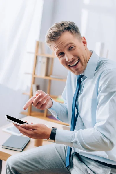 Homme d'affaires joyeux pointant vers smartphone avec écran blanc dans le bureau — Photo de stock