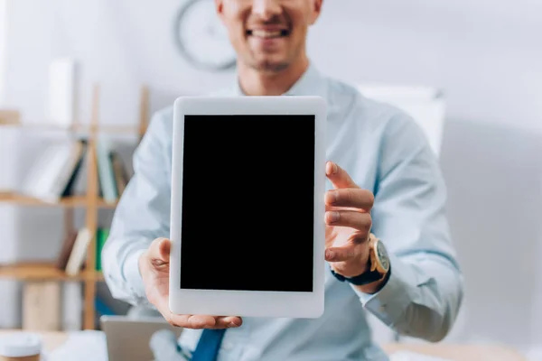 Vista recortada de la tableta digital con pantalla en blanco en manos de un hombre de negocios sonriente sobre un fondo borroso - foto de stock