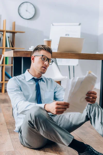 Empresário em óculos olhando para papéis no chão perto da mesa de trabalho — Fotografia de Stock
