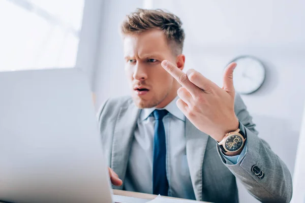 Сердитий бізнесмен показує середній палець під час використання ноутбука на розмитому передньому плані в офісі — стокове фото