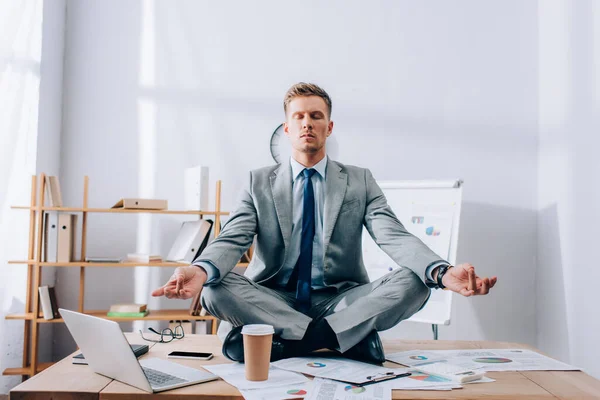 Joven hombre de negocios en traje meditando cerca de papeles y dispositivos en la mesa - foto de stock