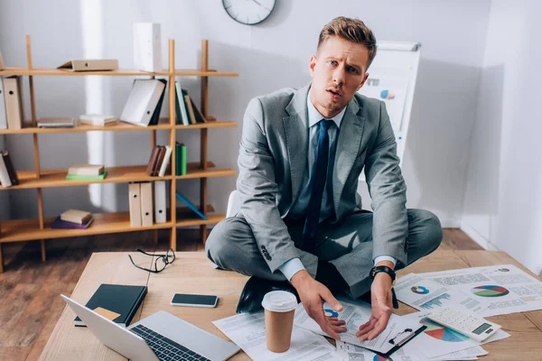 Hombre de negocios confundido mirando la cámara cerca de dispositivos y papeles en la mesa - foto de stock