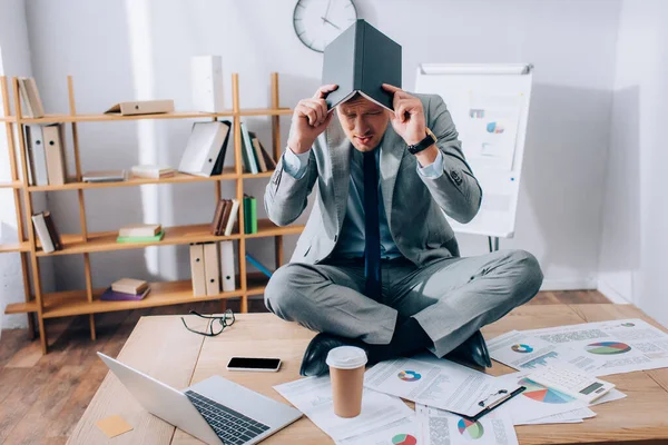 Empresario cubriendo la cabeza con un cuaderno mientras está sentado cerca de la computadora portátil y papeles en la mesa - foto de stock