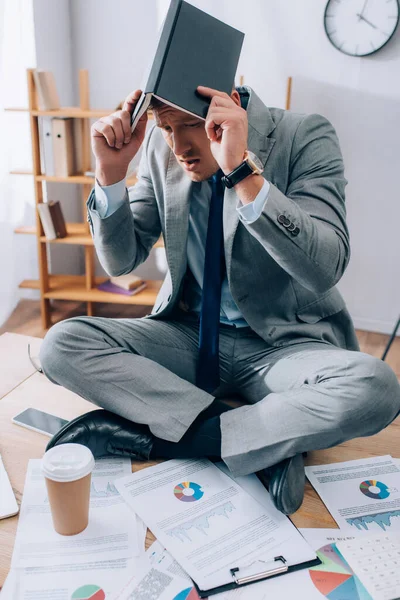 Upset businessman covering head with notebook near documents, smartphone and coffee to go on table — Stock Photo
