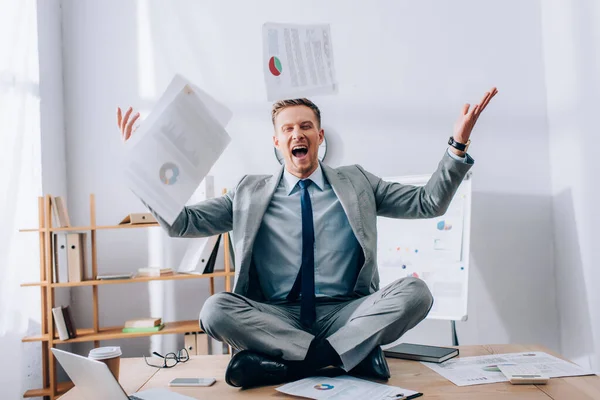 Alegre hombre de negocios lanzando papeles mientras está sentado cerca de dispositivos y café para ir a la mesa - foto de stock