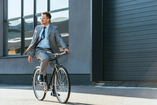 Positive businessman in formal wear and eyeglasses riding bicycle near building — Stock Photo