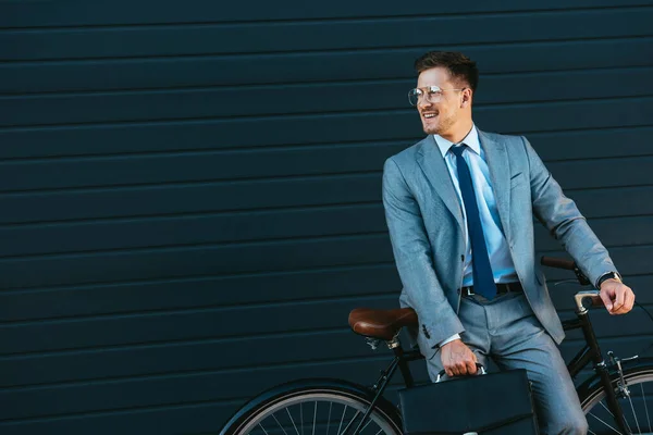 Улыбающийся бизнесмен держит портфель рядом с велосипедом и зданием — стоковое фото