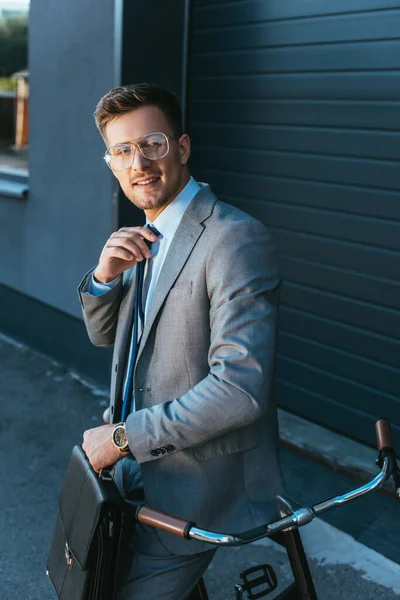 Smiling businessman with briefcase adjusting tie near bike outdoors — Stock Photo