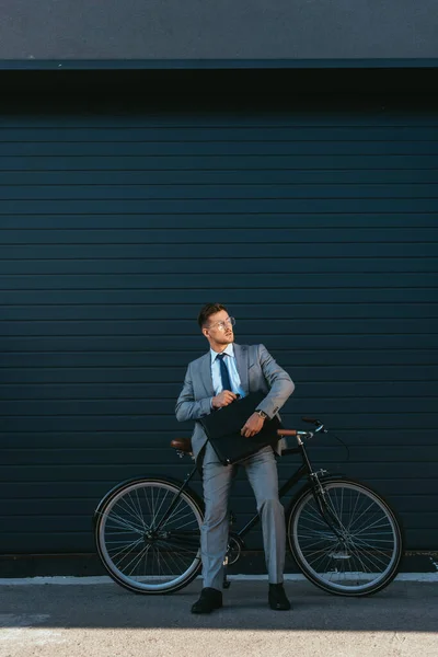 Молодой бизнесмен держит портфель рядом с велосипедом и зданием — стоковое фото