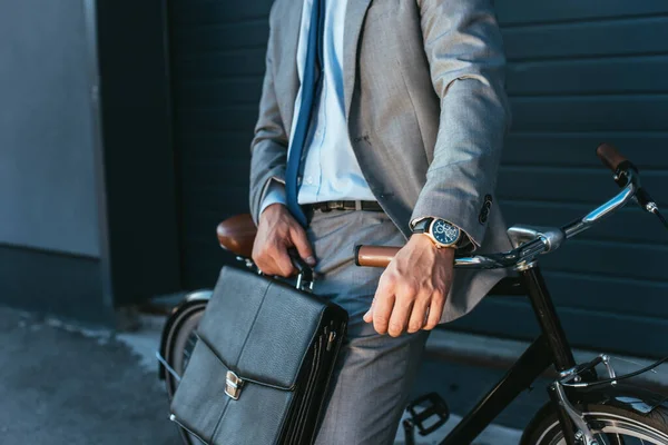 Vista ritagliata dell'uomo d'affari in tuta che tiene la valigetta vicino alla bicicletta all'aperto — Foto stock